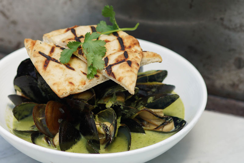 elegantly-plated dish of mussels with green curry topped with naan